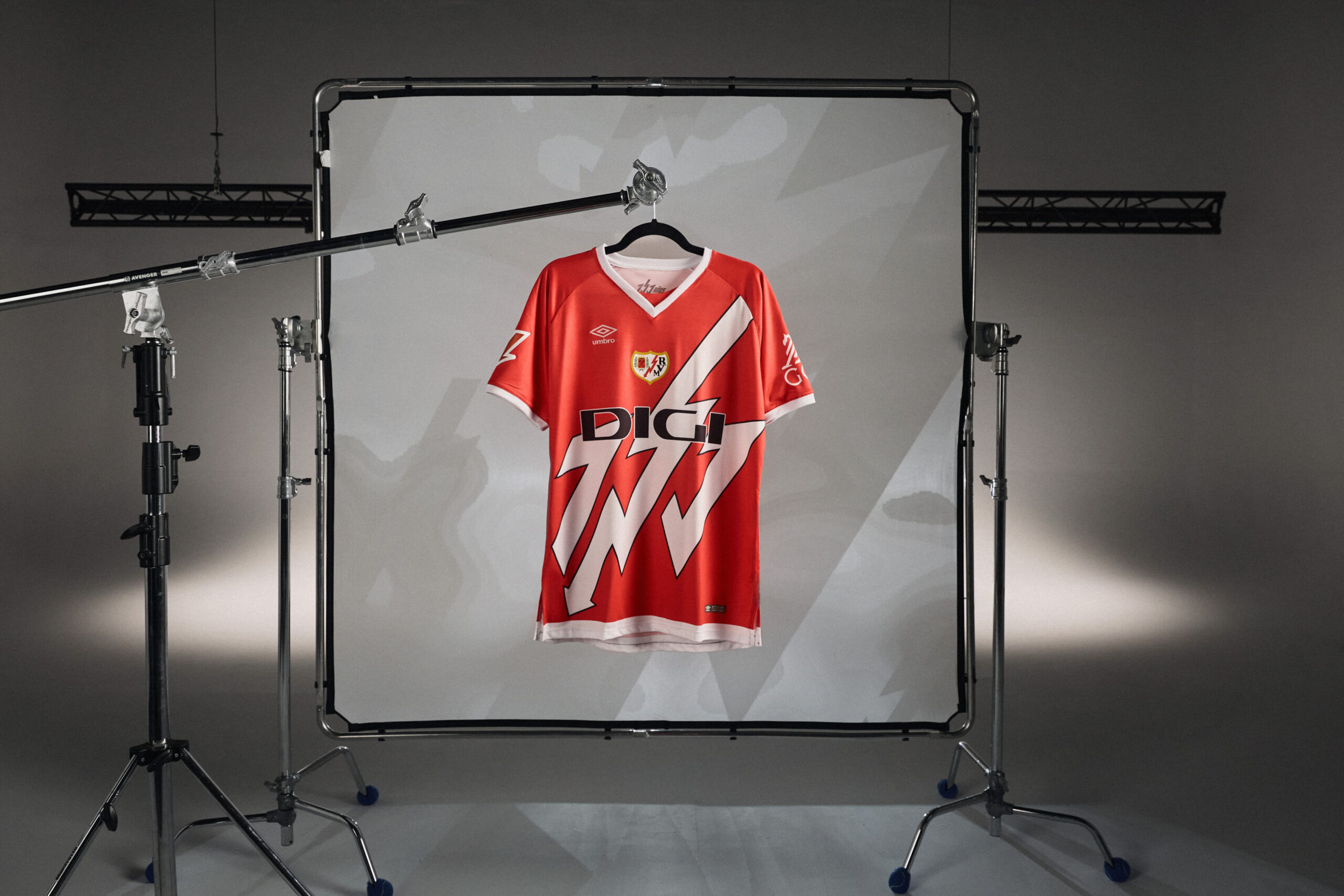 Nueva camiseta de la firma UMBRO para el Rayo Vayecano colgada deuna percha sobre un stand de flas de fotografía cromado con un fondo