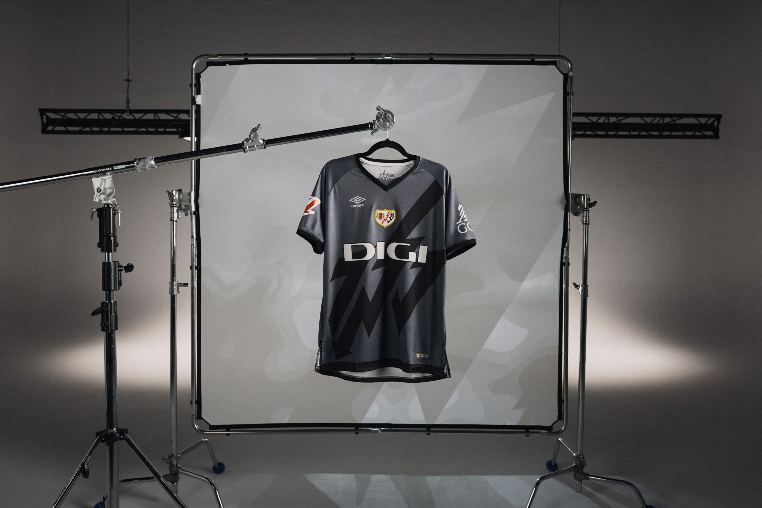 Nueva camiseta de la firma UMBRO para el Rayo Vayecano colgada deuna percha sobre un stand de flas de fotografía cromado con un fondo