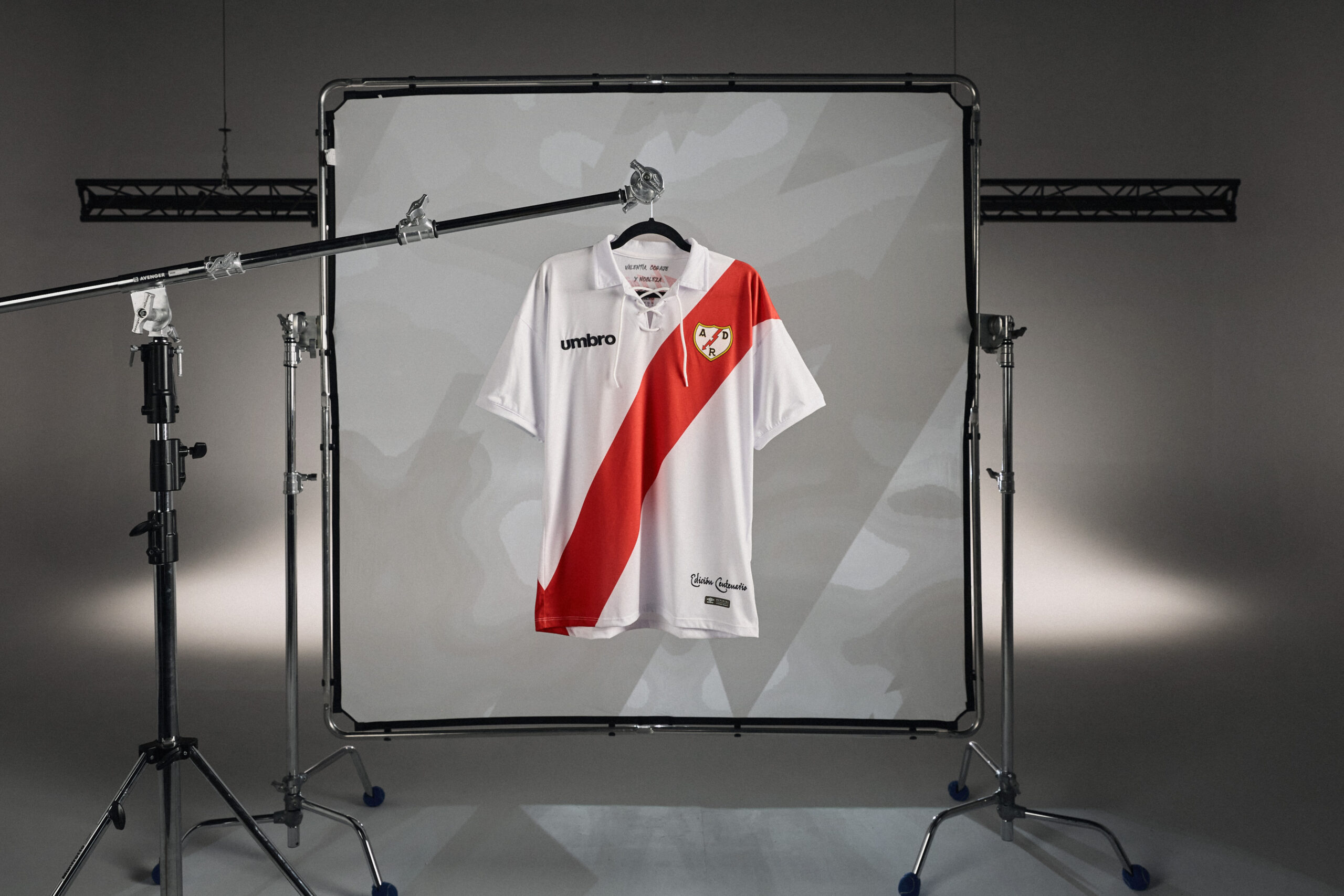 Nueva camiseta de la firma UMBRO para el Rayo Vayecano colgada deuna percha sobre un stand de flas de fotografía cromado con un fondo proyectado con logotipos de la marca